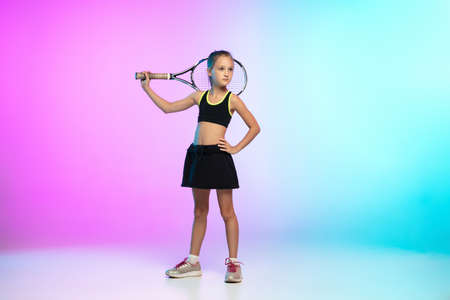Powerful. Little tennis girl in black sportwear isolated on gradient background in neon light. Little caucasian model, sport kid training in motion and action. Sport, movement, childhood concept.の写真素材