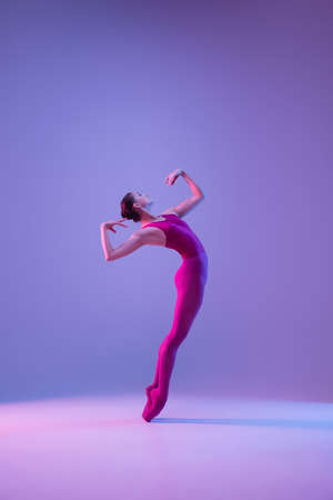 Strong. Young and graceful ballet dancer isolated on purple studio background in neon light. Art, motion, action, flexibility, inspiration concept. Flexible caucasian ballet dancer, weightless jumps.の写真素材