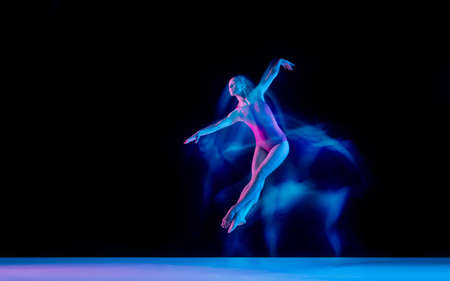 Flying bird. Young and graceful ballet dancer on black studio background in neon mixed light. Art, motion, action, flexibility, inspiration concept. Flexible caucasian ballet dancer, weightless jumps.の写真素材