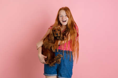 Hugs a dog, happy. Caucasian teen girls portrait on coral pink studio background. Beautiful female redhead model. Concept of human emotions, facial expression, sales, ad, youth culture. Copyspace.の写真素材