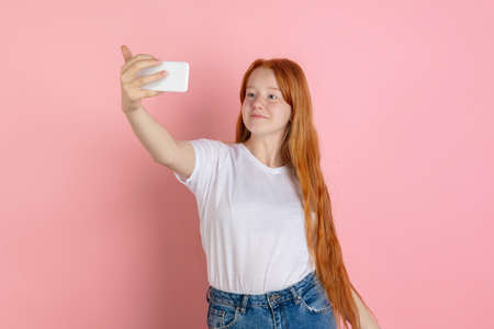 Taking selfie, vlog. Caucasian teen girls portrait on coral pink studio background. Beautiful female redhead model. Concept of human emotions, facial expression, sales, ad, youth culture. Copyspace.の写真素材