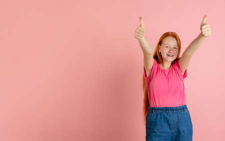 Thumbs up. Caucasian teen girls portrait on coral pink studio background. Beautiful female redhead...