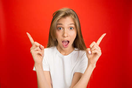 Pointing. Portrait of young caucasian woman with bright emotions on red studio background with copyspace. Blonde female model. Concept of human emotions, facial expression, sales, advertising, youth.の写真素材