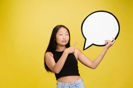 Speech bubble. Portrait of young asian girl isolated on yellow studio background. Stylish, trendy....