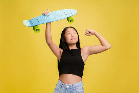 Posing with skate. Portrait of young asian girl isolated on yellow studio background. Stylish,...