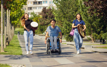 Cheerful. Happy caucasian handicapped man on a wheelchair spending time with friends playing live instrumental music outdoors. Concept of social life, friendship, possibilities, inclusion, diversity.の写真素材