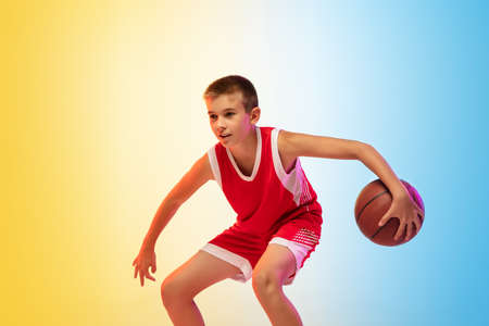 Hurry up. Portrait of young basketball player in uniform on gradient studio background. Teenager confident practicing with ball. Concept of sport, movement, healthy lifestyle, ad, action, motion.の写真素材