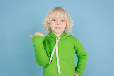 Pointing at side. Portrait of beautiful caucasian little boy isolated on blue studio background. Blonde curly male model. Concept of facial expression, human emotions, childhood, ad, sales. Copyspace.の写真素材