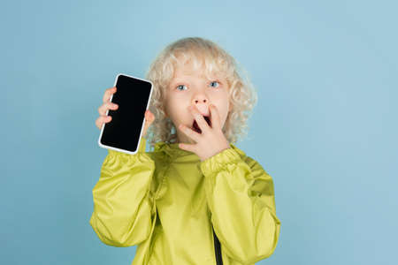 Showing phone screen. Portrait of beautiful caucasian little boy isolated on blue studio background....