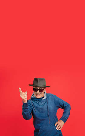 Pointing. Portrait of senior man in stylish eyewear and hat isolated on red studio background. Tech and joyful elderly lifestyle concept. Trendy colors, forever youth. Copyspace for your ad. Flyerの写真素材