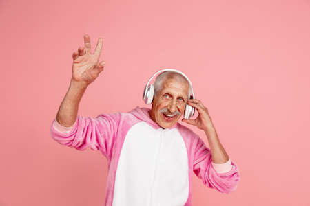 Pointing up in headphones. Portrait of senior hipster man in stylish attire isolated on pink studio background. Tech and joyful elderly lifestyle concept. Trendy colors, forever youth. Copyspaceの写真素材