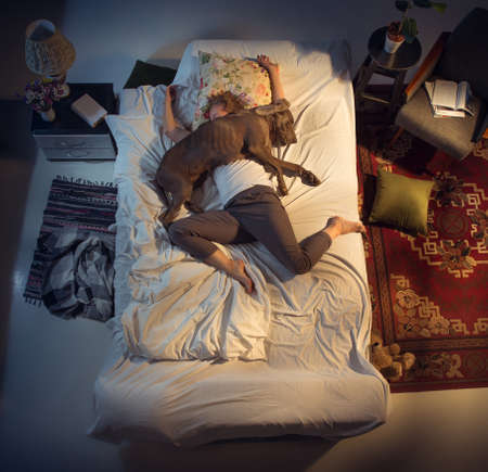 Portrait of a woman, female breeder sleeping in the bed with her dog at home. Top view. Dressed housekeeper sleeping after tiring work day. Holding his pet close to. Job, work, pets love concept.の写真素材