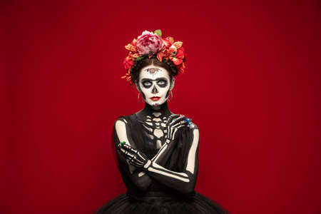 Gambling. Young girl like Santa Muerte Saint death or Sugar skull with bright make-up. Portrait isolated on red studio background with copyspace. Celebrating Halloween or Day of the dead.の写真素材