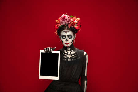 Holding tablet. Young girl like Santa Muerte Saint death or Sugar skull with bright make-up. Portrait isolated on red studio background. Celebrating Halloween or Day of the dead. Copyspace on screen.の写真素材