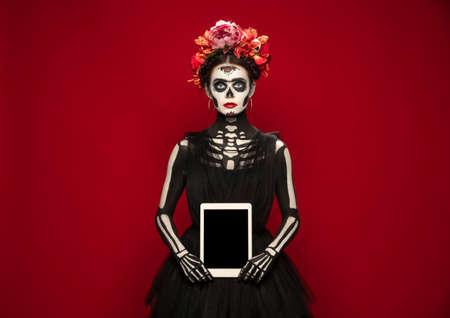 Holding tablet. Young girl like Santa Muerte Saint death or Sugar skull with bright make-up. Portrait isolated on red studio background. Celebrating Halloween or Day of the dead. Copyspace on screen.の写真素材