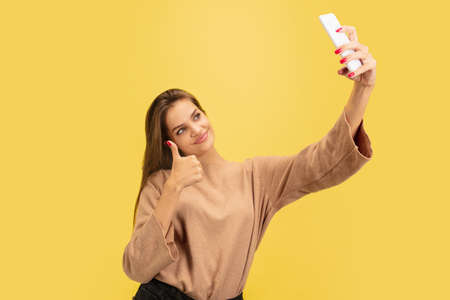 Taking selfie. Portrait of young caucasian woman isolated on yellow studio background with...