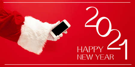 Hand of Santa Claus holding phone, greeting with New Year 2021 and Christmas. Red background with...