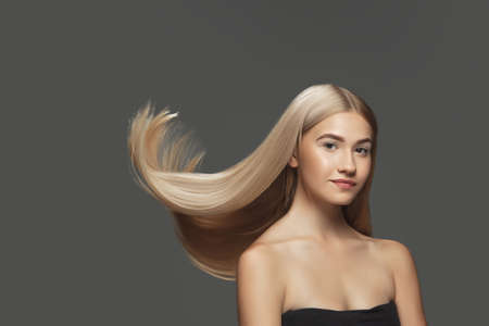 Youth. Beautiful model with long smooth, flying blonde hair on dark grey studio background. Young caucasian model with well-kept skin and hair blowing on air. Concept of salon care, beauty, fashion.の写真素材