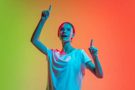 Pointing up. Young caucasian girls portrait on gradient green-orange studio background in neon light. Concept of youth, human emotions, facial expression, sales, advertising. Beautiful teen model.の写真素材