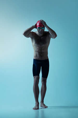 Professional caucasian male swimmer with hat and goggles practicing and training isolated on blue neoned studio background. Grace of motion and action. Healthy lifestyle, sport and movement concept.の写真素材