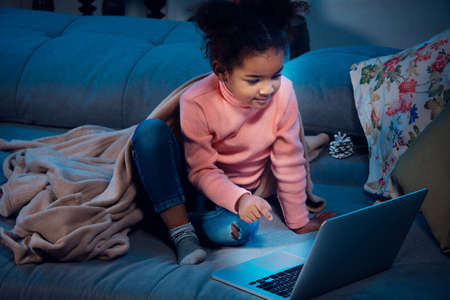 Happy african-american little girl during video call with laptop and home devices, looks delighted...
