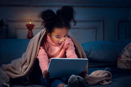 Happy african-american little girl during video call with laptop and home devices, looks delighted...