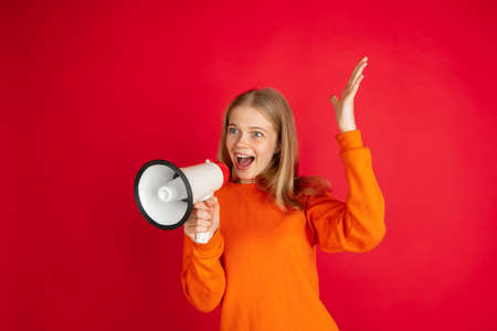 Shouting with megaphone, loudspeaker. Portrait of young woman isolated on red studio background with copyspace. Beautiful female model. Concept of human emotions, facial expression, sales, ad, youth.の写真素材