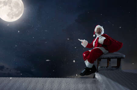Emotional Santa Claus congratulating with New Year 2021 and Christmas. Man in traditional costume sitting on house roof with full moon on background in midnight. Winter, holidays, sales. Copyspace.の写真素材