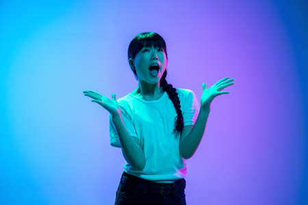 Shocked, astonished. Young asian womans portrait on gradient blue-purple studio background in neon light. Concept of youth, human emotions, facial expression, sales, ad. Beautiful brunette model.の写真素材