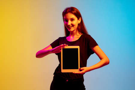 Showing blank tablet screen. Young caucasian womans portrait on gradient blue-yellow studio background in neon light. Concept of youth, human emotions, facial expression, sales, ad. Beautiful model.の写真素材