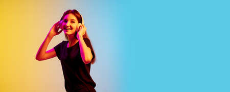Listening to music with headphones. Young caucasian womans portrait on gradient blue-yellow studio...
