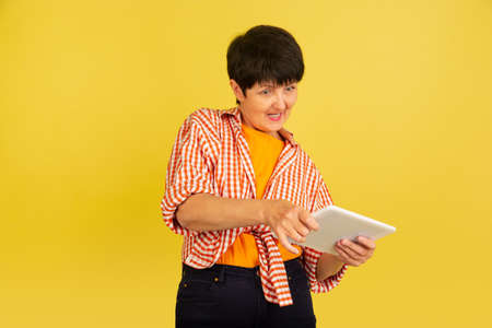 Playing with tablet. Portrait of senior woman in stylish outfit isolated on yellow studio...
