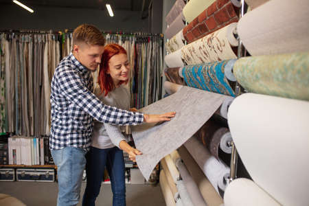 Cover. Couple choosing textile at home decoration store, shop. Making of home interior design during quarantine. Happy man and woman, young family look dreamful, cheerful choosing materials.の写真素材
