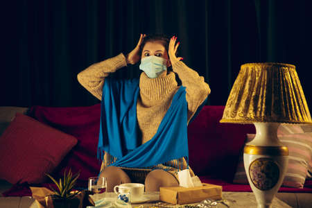 Woman wrapped in a plaid wearing face mask trying to protect herself from somebodys sick, looks disguasted, angry, sad sitting on sofa at home indoors. Healthcare and medicine, ill prevention.の写真素材