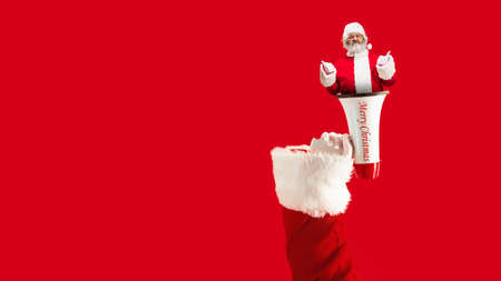 Greeting flyer for ad. Concept of Christmas, 2021 New Years party, winter mood, holidays. Copyspace, postcard. Big hand with loudspeaker, megaphone with Santa Claus inside it on red backgroundの写真素材
