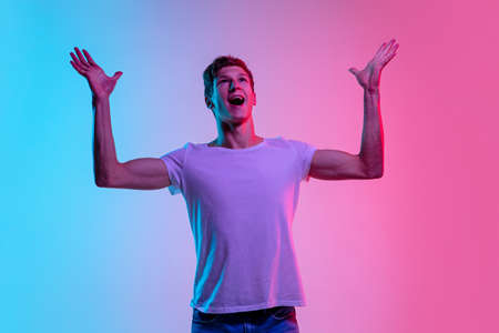 Astonished. Young caucasian mans portrait on gradient blue-pink studio background in neon light. Concept of youth, human emotions, facial expression, sales, ad. Beautiful model in casual.の写真素材