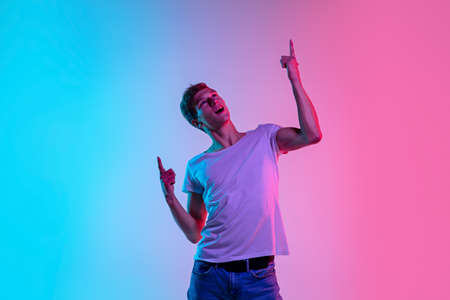 Pointing up. Young caucasian mans portrait on gradient blue-pink studio background in neon light. Concept of youth, human emotions, facial expression, sales, ad. Beautiful model in casual.の写真素材