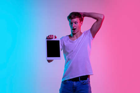 Shocked with tablet. Young caucasian mans portrait on gradient blue-pink studio background in neon light. Concept of youth, human emotions, facial expression, sales, ad. Beautiful model in casual.の写真素材