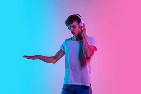 Music in headphones. Young caucasian mans portrait on gradient blue-pink studio background in neon light. Concept of youth, human emotions, facial expression, sales, ad. Beautiful model in casual.の写真素材
