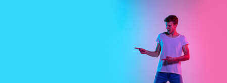 Pointing at side, flyer. Young caucasian mans portrait on gradient blue-pink studio background in neon light. Concept of youth, human emotions, facial expression, sales, ad. Beautiful model in casual.の写真素材