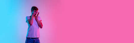 Scared, flyer. Young caucasian mans portrait on gradient blue-pink studio background in neon light. Concept of youth, human emotions, facial expression, sales, ad. Beautiful model in casual.の写真素材