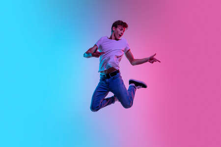 Pointing at side. Young caucasian mans jumping high on gradient blue-pink studio background in neon light. Concept of youth, human emotions, facial expression, sales, ad. Beautiful model in casual.の写真素材