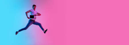 Jumping high with tablet, flyer. Young mans portrait on gradient blue-pink studio background in neon light. Concept of youth, human emotions, facial expression, sales, ad. Beautiful model in casual.の写真素材