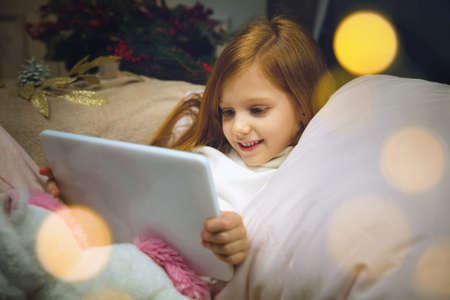 Dreams. Happy caucasian little girl during video call with laptop and home devices, looks dreamful and happy. Talking to Santa before New Years eve, her family, watching cartoons, typing text. Bokehの写真素材