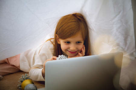 Wishes. Happy caucasian little girl during video call with laptop and home devices, looks dreamful and happy. Talking to Santa before New Years eve, her family, watching cartoons, typing text. Bokehの写真素材