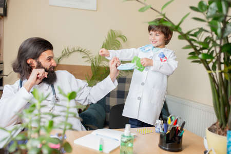 Paediatrician doctor examining a child in comfortabe medical office. Healthcare, childhood, medicine, protection and prevention concept. Little boy trust to doctor. Having fun while wearing face maskの写真素材