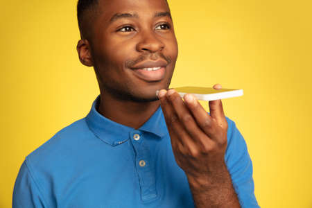 Recording voice message. Young african-american mans portrait isolated on yellow studio background, facial expression. Beautiful male model, copyspace. Concept of human emotions, sales, ad.の写真素材