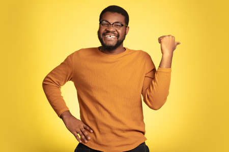 Pointing at side. Young african-american mans portrait isolated on yellow studio background, facial expression. Beautiful male model, monochrome, copyspace. Concept of human emotions, sales, ad.の写真素材