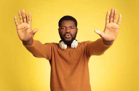 Stopping. Young african-american mans portrait isolated on yellow studio background, facial expression. Beautiful male model with headphones, copyspace. Concept of human emotions, sales, ad.の写真素材
