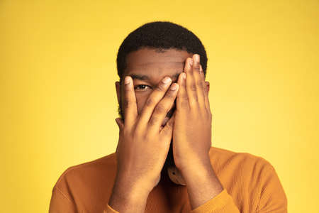Scared hiding face. Young african-american mans portrait isolated on yellow studio background, facial expression. Beautiful male model with headphones, copyspace. Concept of human emotions, sales, ad.の写真素材